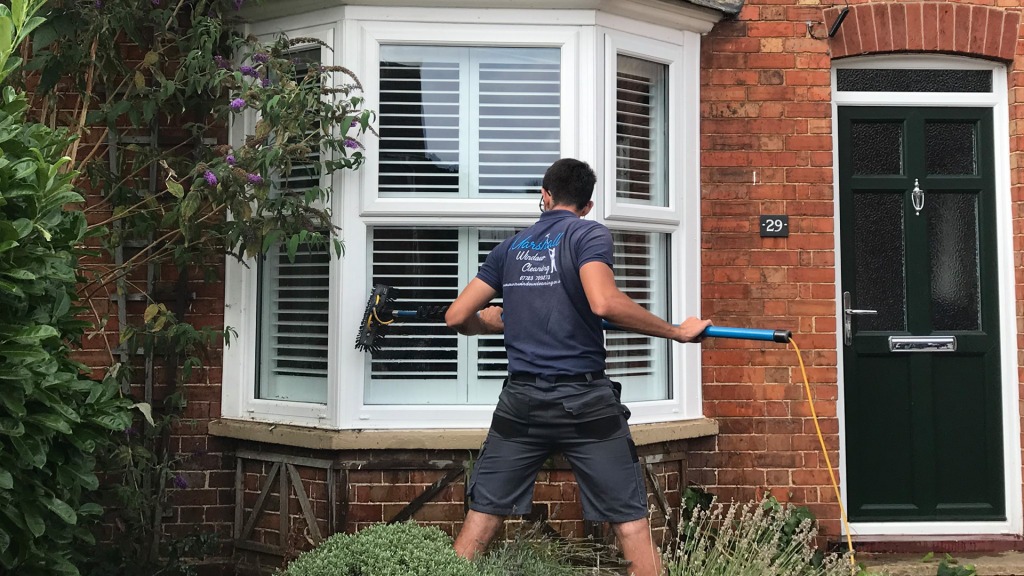 Professional Marshall Window Cleaning technician in uniform using Reach & Wash water-fed pole system to clean bay windows on a red brick house